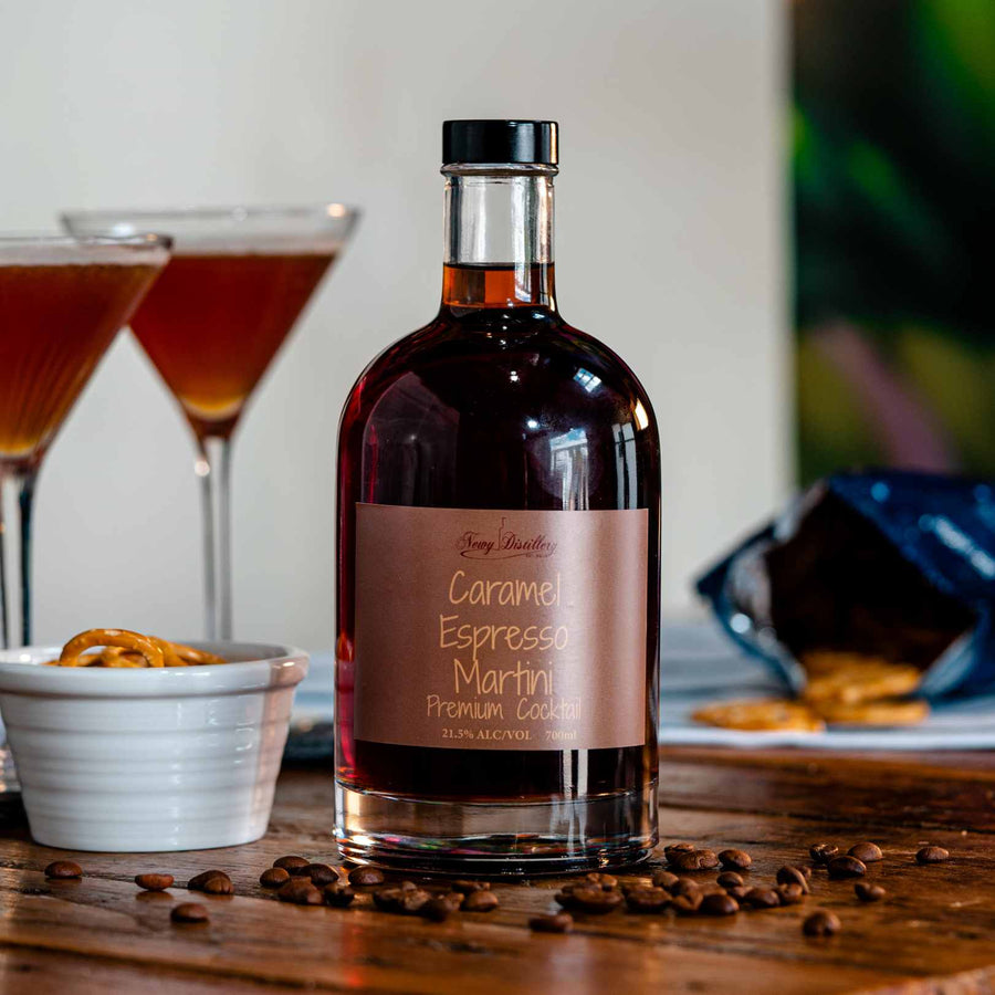 Newy Distillery Caramel Espresso Martini Bottle 700ml. Displayed on brown wooden tablen next to small pot of pretzels and scattering of coffee beans. Two Caramel Espresso Martini cocktails behind, with bag of pretzels on blue and white cloth in the background.