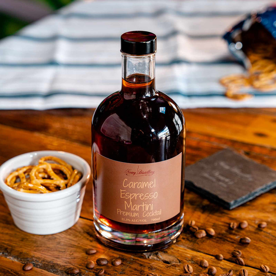 Newy Distillery Caramel Espresso Martini Bottle 700ml. Displayed on brown wooden tablen next to small pot of pretzels and scattering of coffee beans. Bag of pretzels on blue and white cloth in the background.