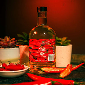 Chilli Gin by Newy Distillery. Award winning dry gin infiused with fresh chillis. 500ml bottle displayed on grey slate surroudn by cayenne and birdseye chillis.