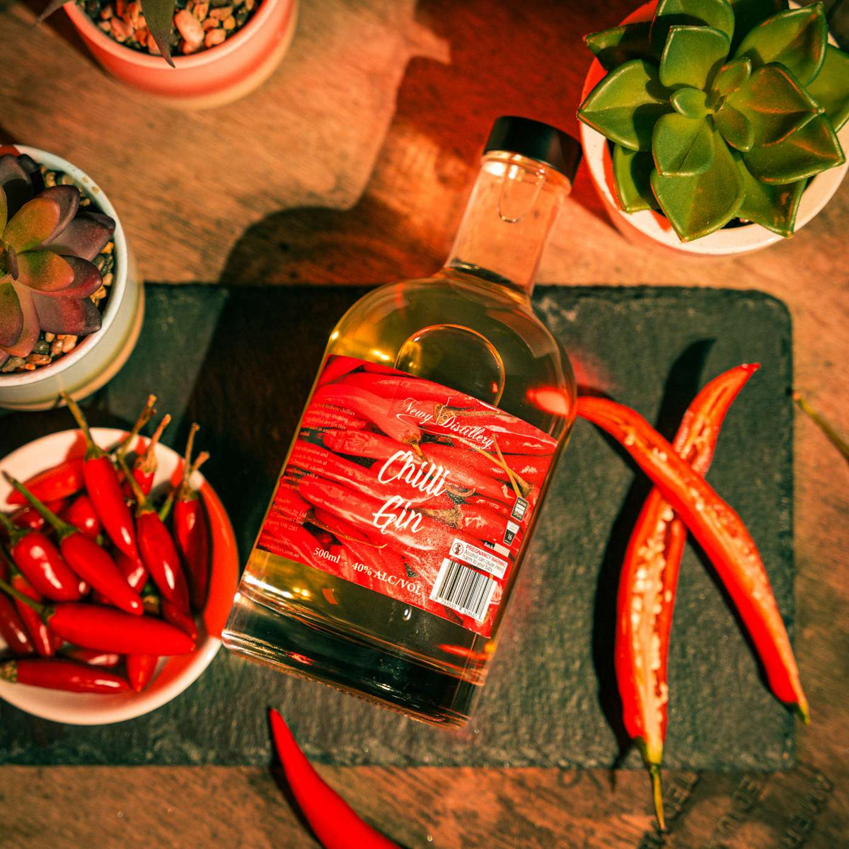 Chilli Gin by Newy Distillery. Award winning dry gin infiused with fresh chillis. 500ml bottle displayed on grey slate surroudn by cayenne and birdseye chillis.