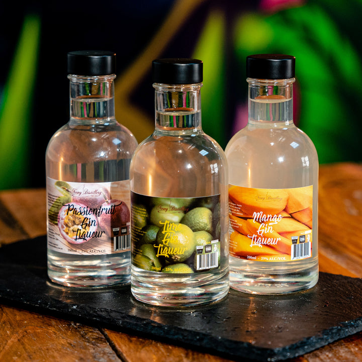 Fruit Gin Liqueur Gift Pack. 3 gin liqueurs gift set. 200ml bottle lime gin liqueur. 200ml bottle mango gin liqueur, 200ml bottle passionfruit gin liqueur. Three bottles on grey slate with colourful background. By Newy Distillery.