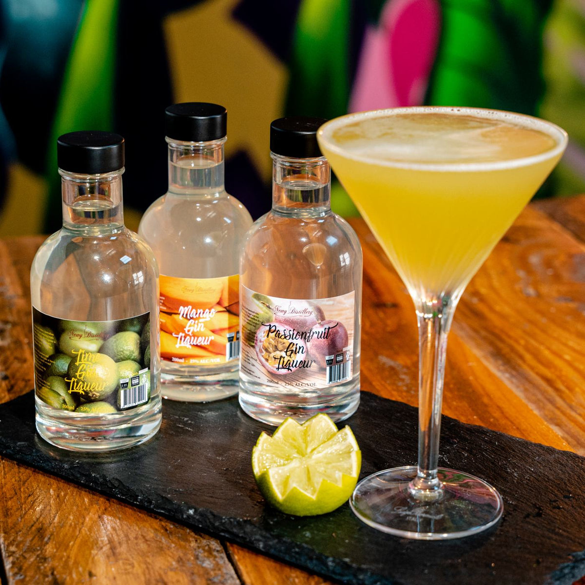 Fruit Gin Liqueur Gift Pack. 3 gin liqueurs gift set. 200ml bottle lime gin liqueur. 200ml bottle mango gin liqueur, 200ml bottle passionfruit gin liqueur. Three bottles on grey slate with colourful background with cocktail in martini glass and lime decoration. By Newy Distillery.