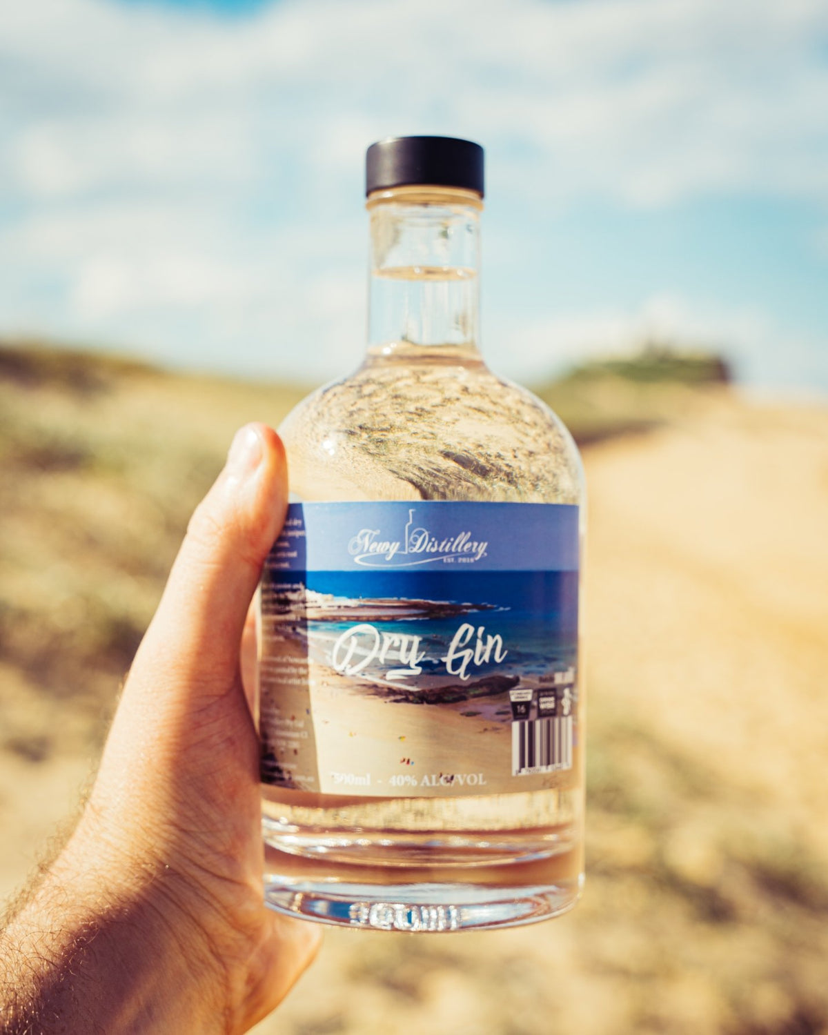 Newy Distillery multi-award Dry Gin 500ml bottle. John Earle label  held in a hand at the beach.